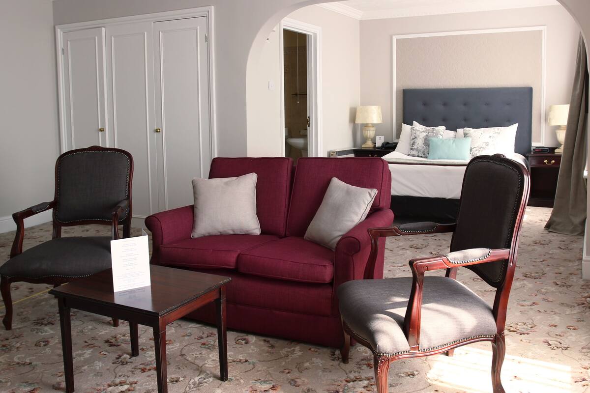 A hotel room with a red sofa, two armchairs, a wooden coffee table, and a bed with a blue headboard.