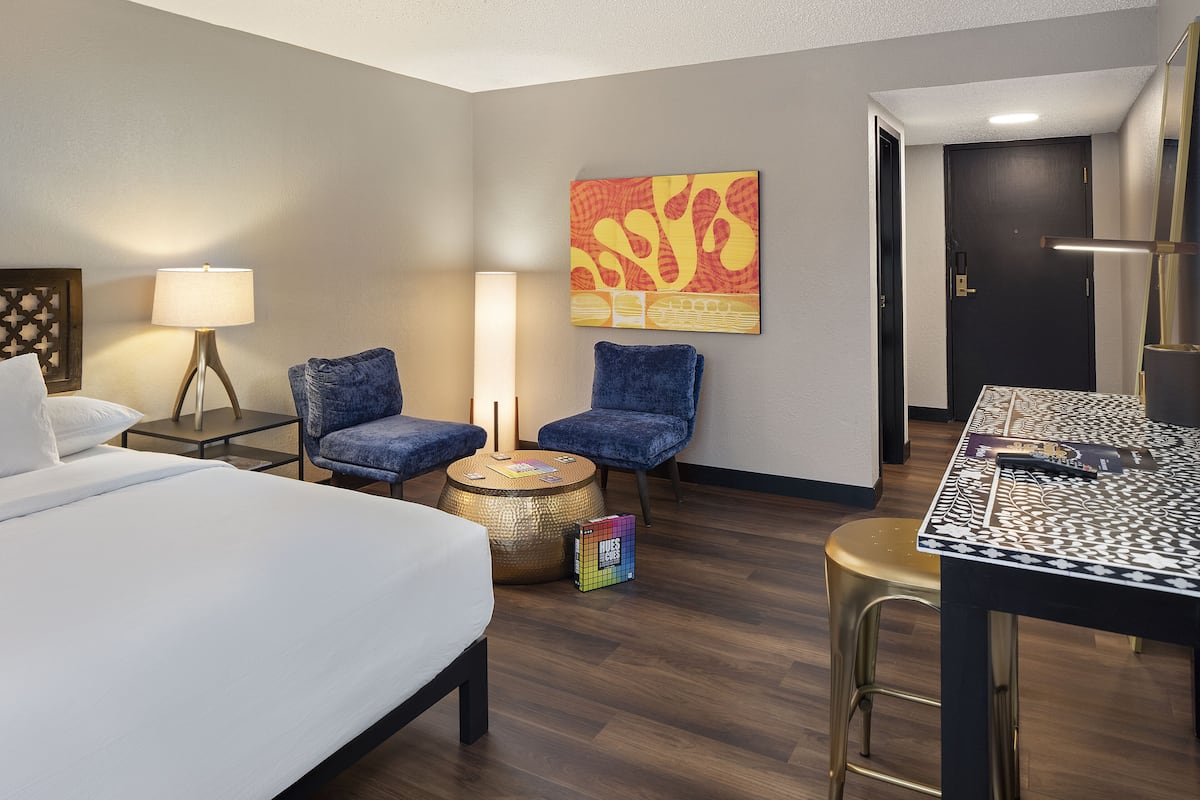Une chambre d’hôtel avec un lit, deux fauteuils, une petite table ronde, une lampe et un tableau au mur.