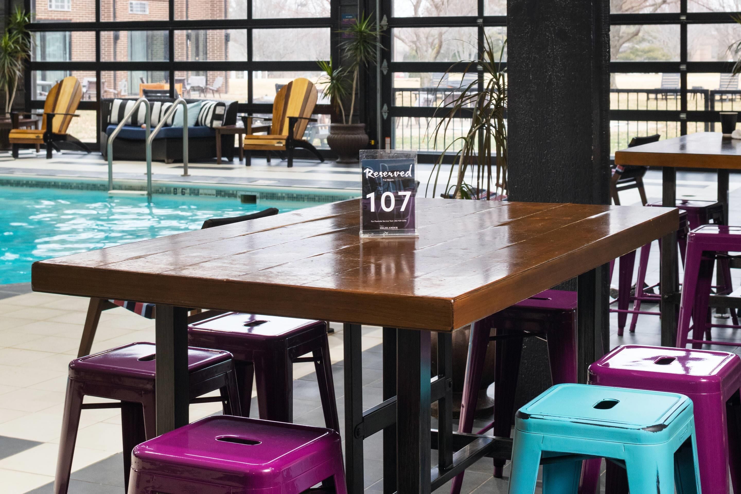 A poolside area with a wooden table and colorful stools, a 'Reserved 107' sign, and lounge chairs in the background.