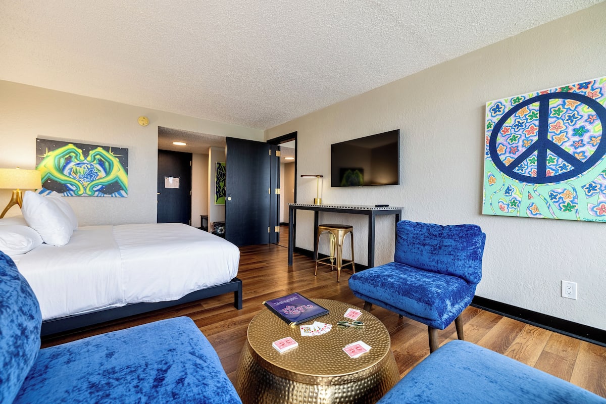 Une chambre d’hôtel avec un lit, deux fauteuils bleus, une table basse ronde, un bureau et un téléviseur fixé au mur.