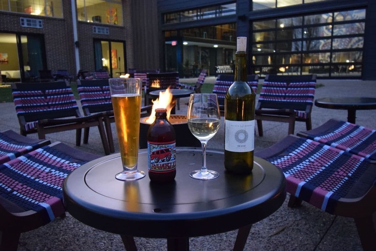 A table with a bottle of wine, a glass of white wine, and a tall glass of beer, surrounded by lounge chairs with patterned cushions.