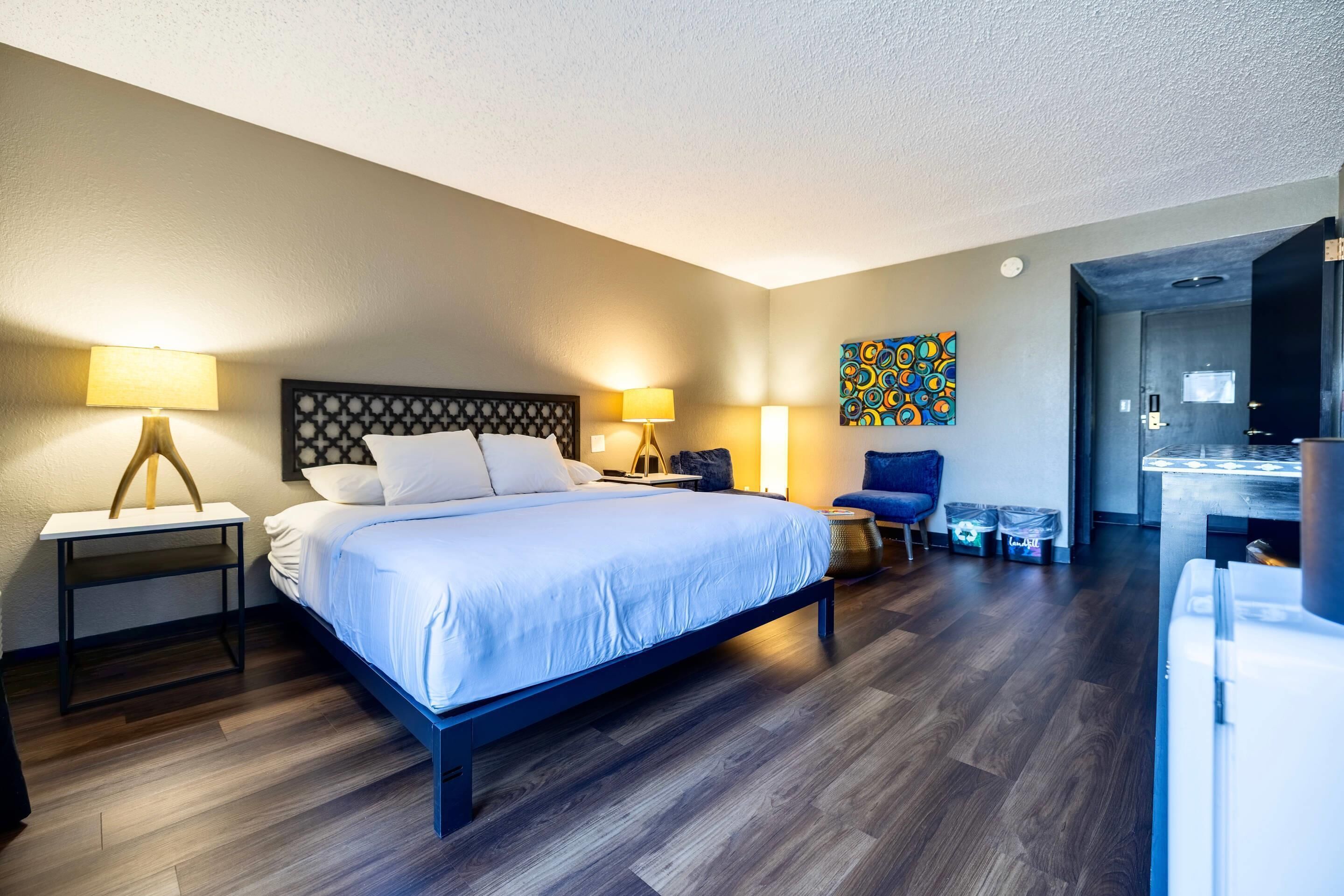 A hotel room with a bed, bedside tables, a chair, a desk, and a refrigerator.