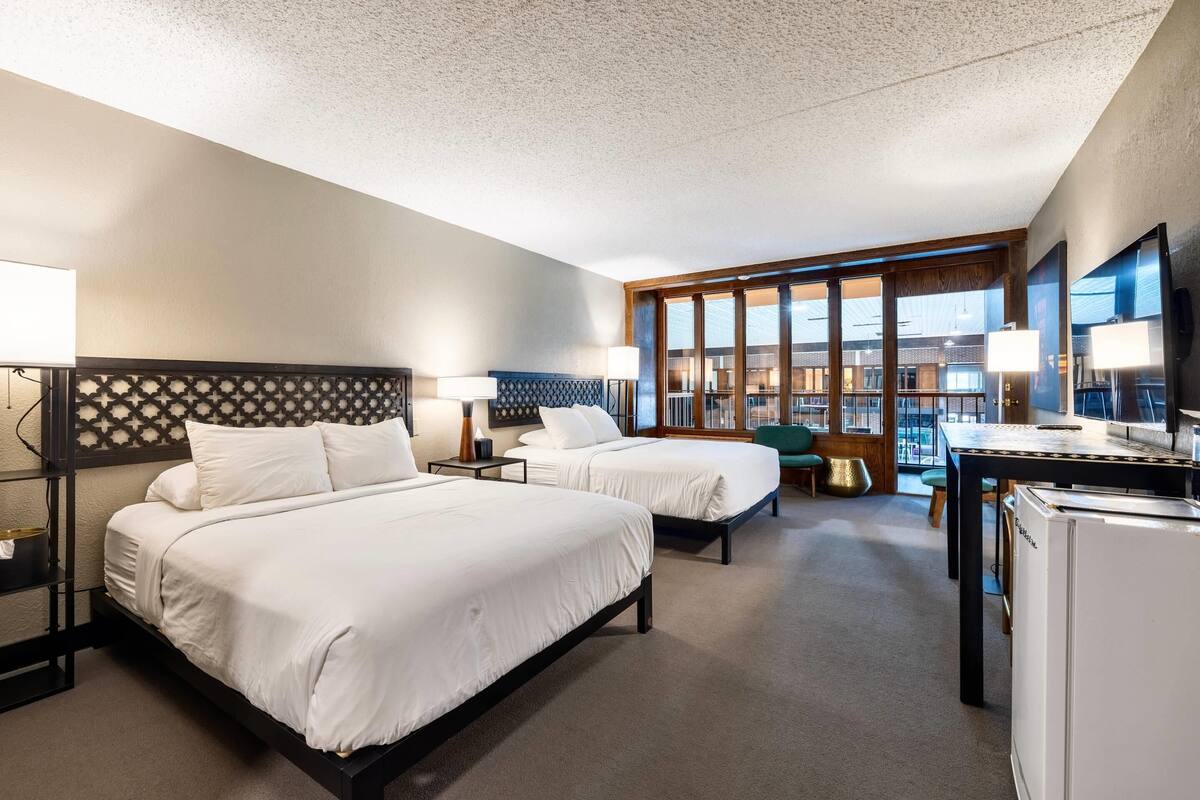Une chambre d’hôtel avec deux lits, un bureau, un r éfrigérateur et une vue sur le paysage urbain grâce à de grandes fenêtres.