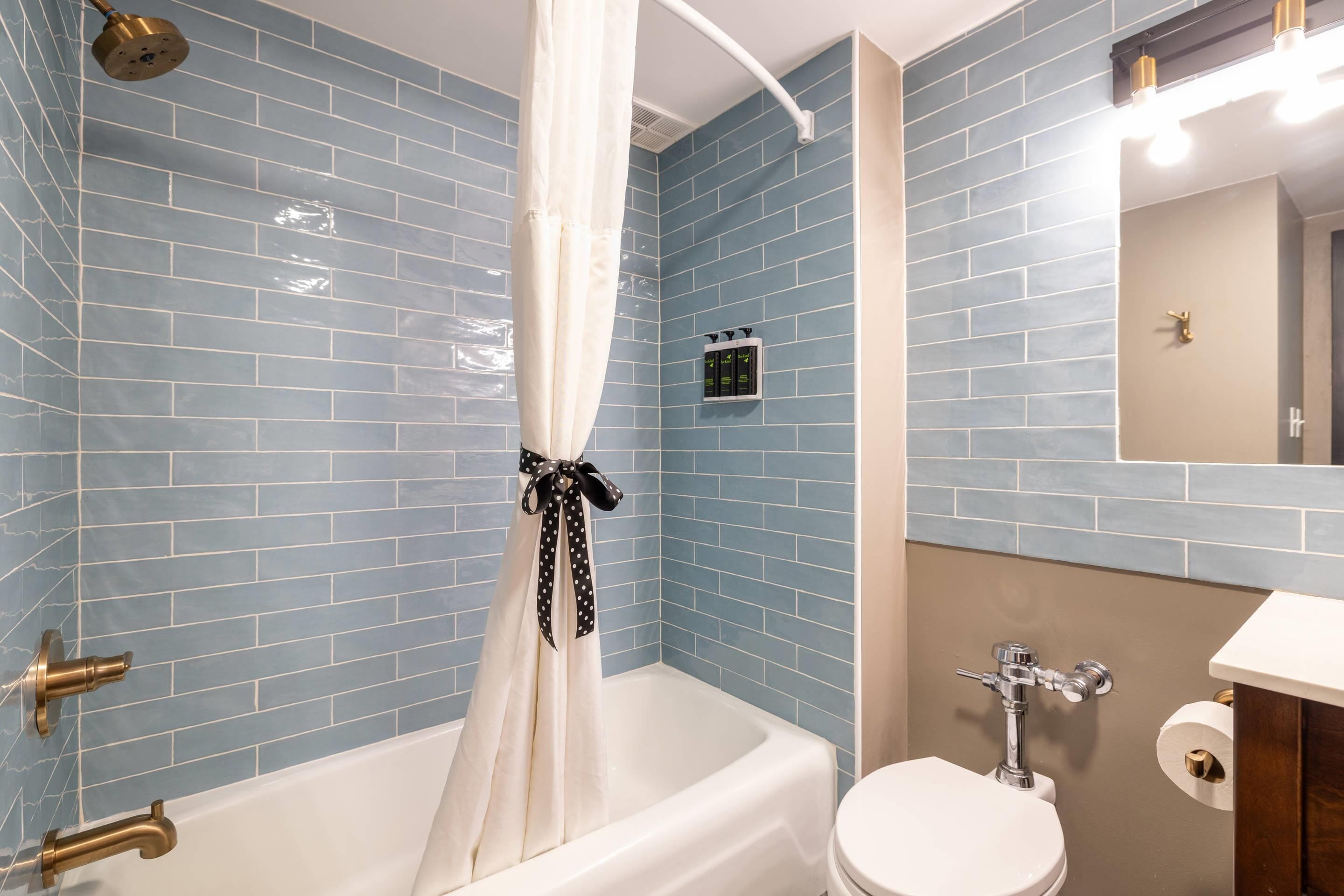 Une salle de bain avec des murs recouverts de carreaux bleus, une baignoire blanche avec un rideau, des toilettes et un miroir.