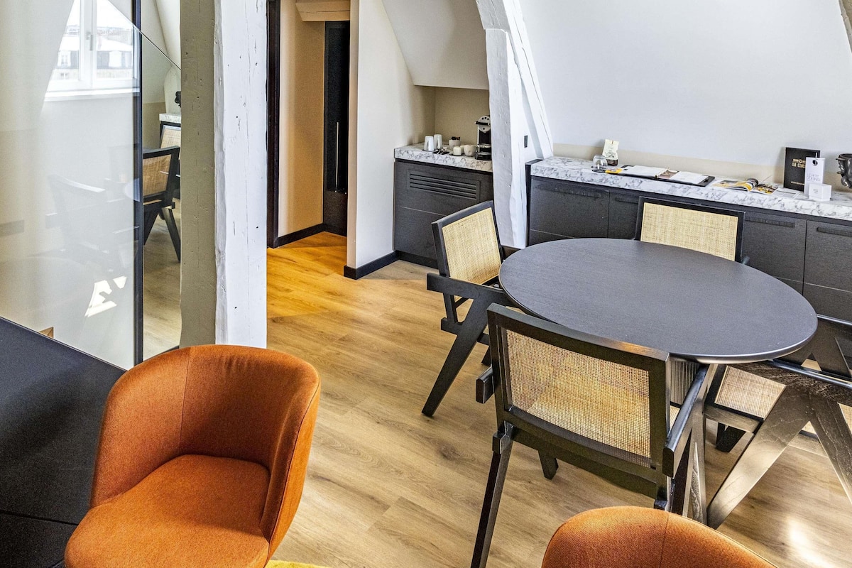 A modern loft-style room with a dining table, two orange chairs, a kitchen area, and a view through a glass door.