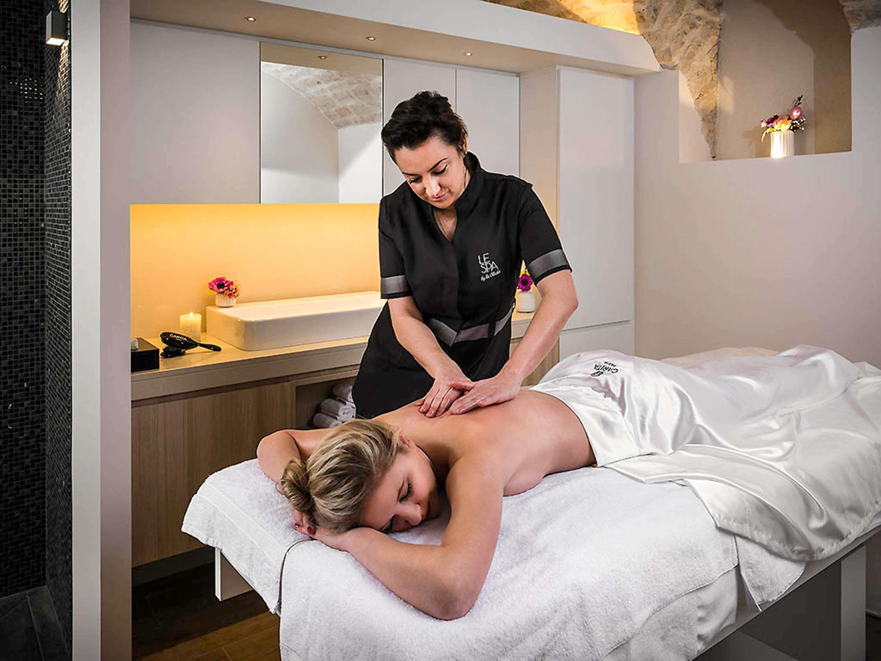 Sesión de masoterapia en un spa, con un cliente recostado en una camilla de tratamiento, un terapeuta atendiendo y una zona de recepción con mostrador y un jarrón de flores.
