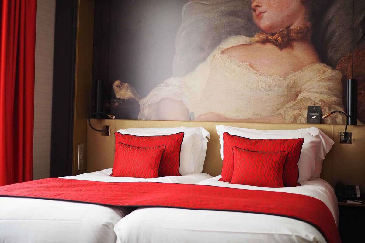 Ein Hotelzimmer mit einem großen Bett, rotem und weißem Bettzeug, einem Gemälde über dem Bett und einem roten Vorhang links.