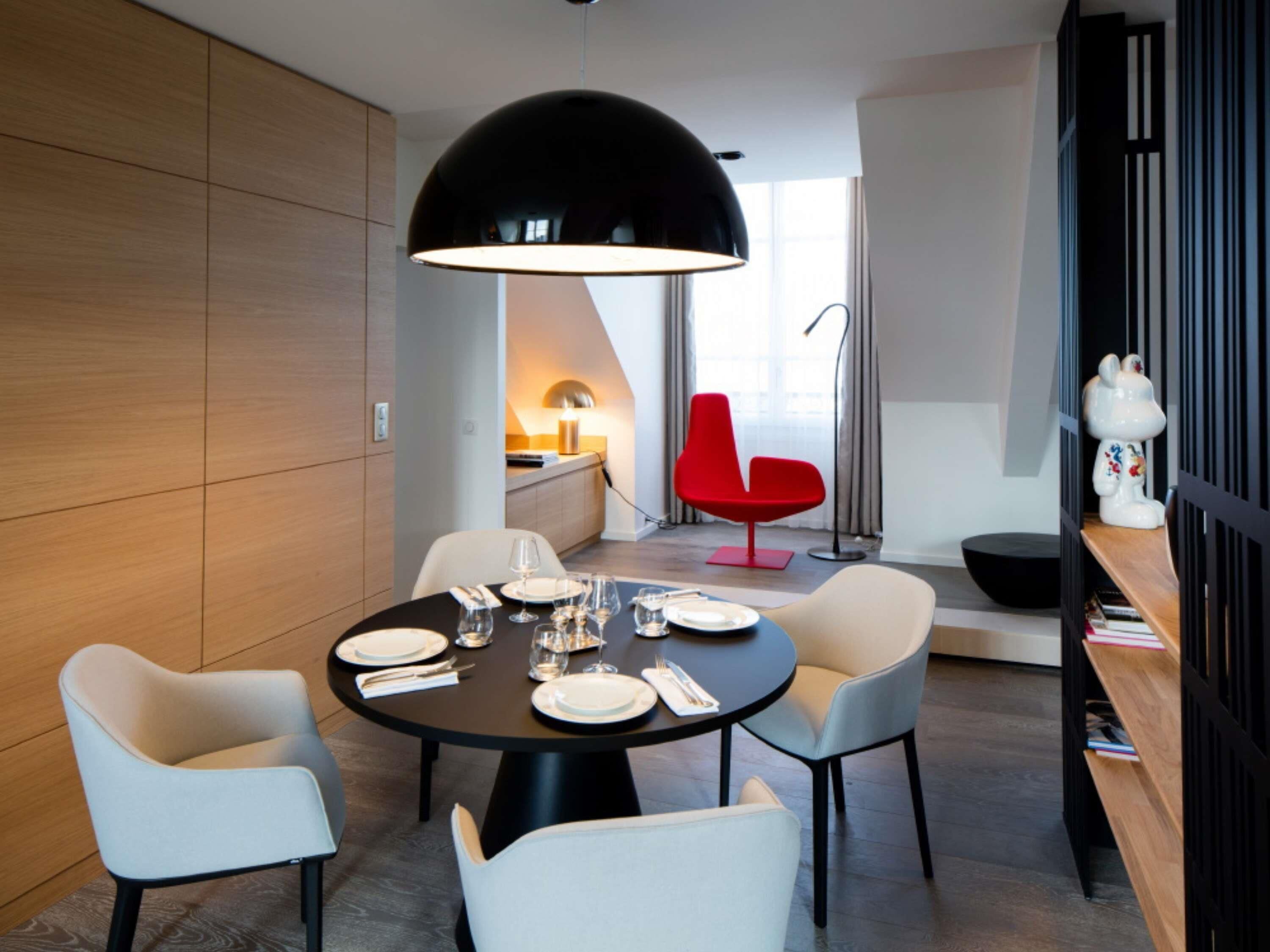 Un comedor moderno con una mesa redonda, cuatro sillas, una lámpara colgante negra, una silla roja y una estantería con objetos decorativos.