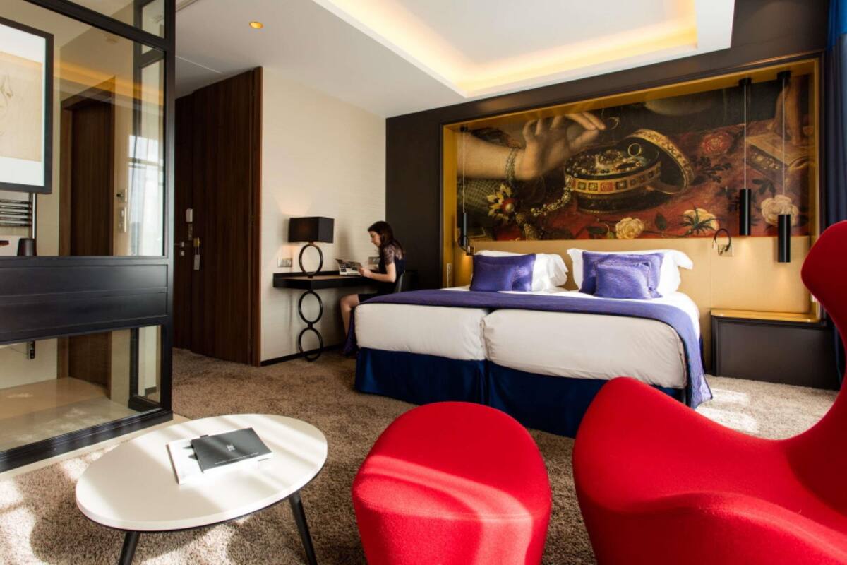 Ein Hotelzimmer mit einem Bett, einem Schreibtisch, einem Laptop, einem roten Sessel und einem großen Gemälde an der Wand.