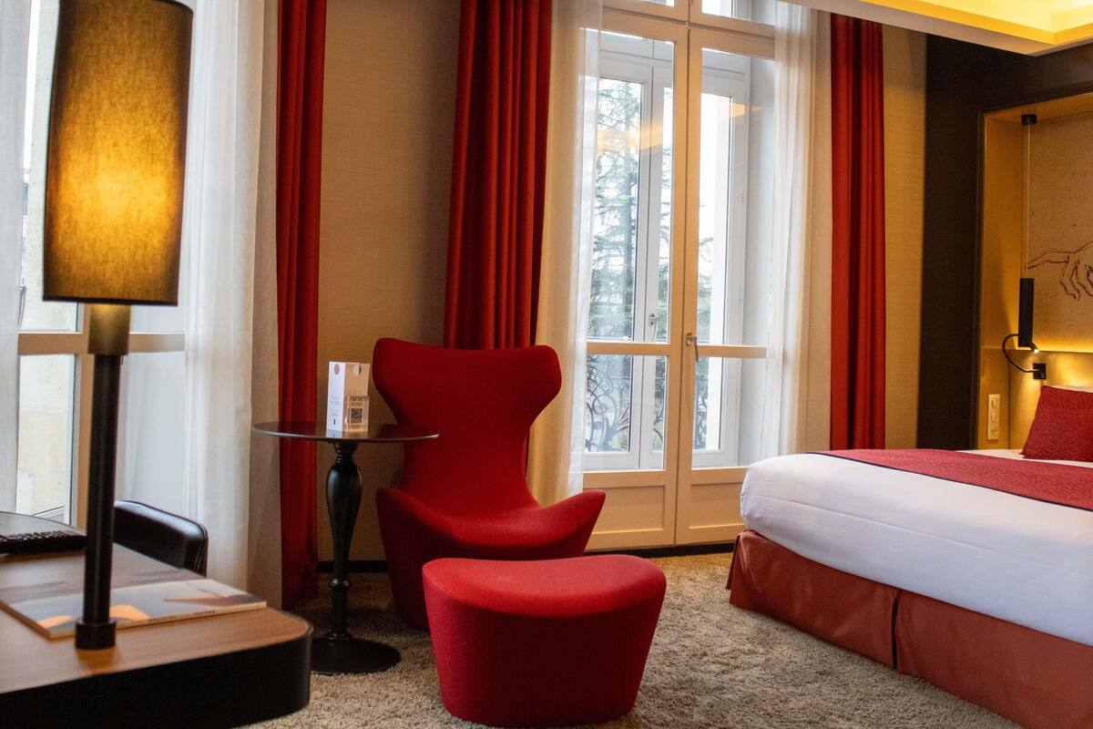 Ein Hotelzimmer mit Bett, Schreibtisch mit Lampe, Stuhl und einem Fenster mit Vorhängen.