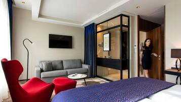 A hotel room with a bed, a sofa, a red chair, a coffee table, a desk, and a bathroom with a glass door.