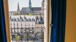 Ein Fenster mit einem Metallbalkongeländer, Blick auf eine Stadtlandschaft mit Gebäuden und einem Kirchturm.