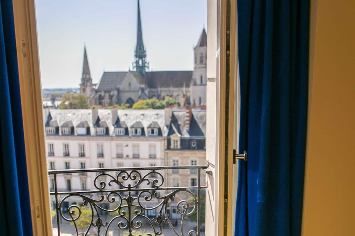 Une fenêtre avec un balcon en métal, d’où l’on peut admirer un paysage urbain avec des immeubles et le clocher d’une église.