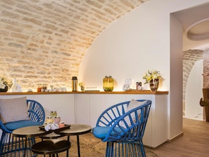 A cozy interior with a stone wall, a curved white ceiling, a wooden counter with jars and a camera, and two blue chairs. - Grand Hôtel La Cloche Dijon - MGallery Collection (Dijon)