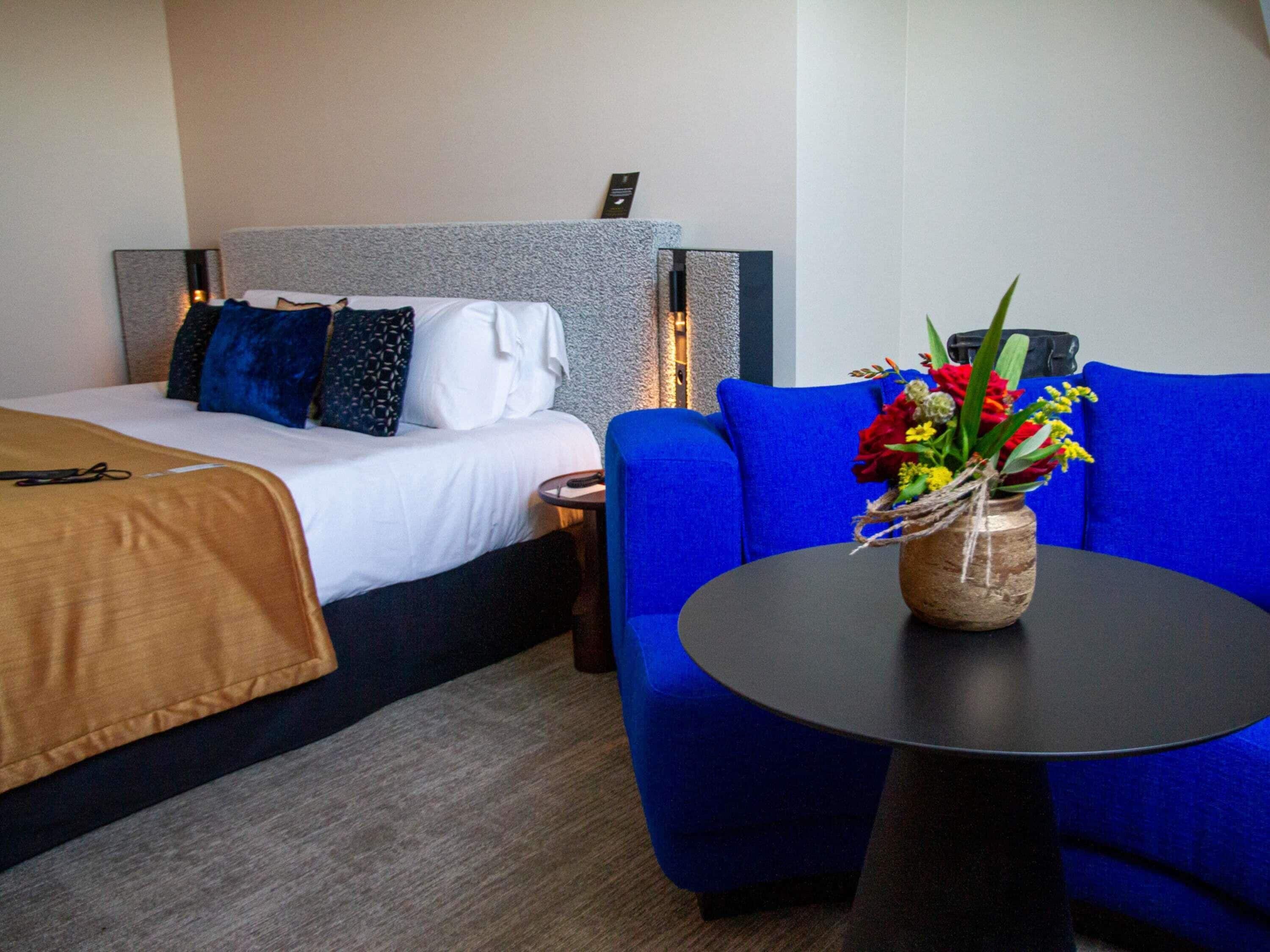 A hotel room with a bed, a blue sofa, a small table with a vase of flowers, and a headboard with built-in lighting.