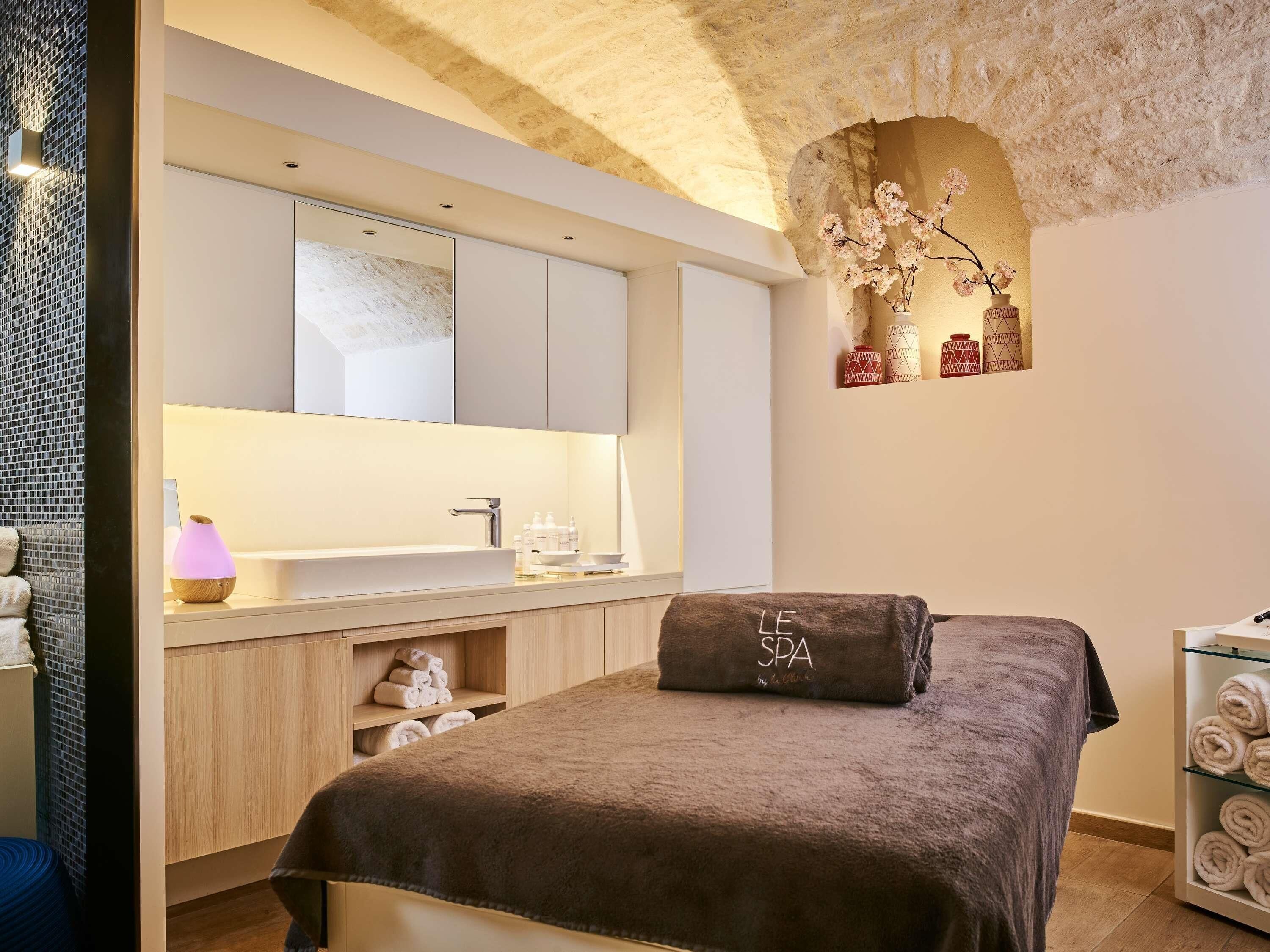 A modern spa room with a large bed, a vanity with a mirror, a sink, and a towel rack.