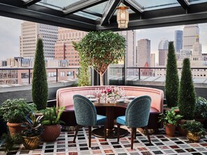 A rooftop dining area with a round table set for two, surrounded by plush seating and potted plants. - The Newbury Boston (Boston)