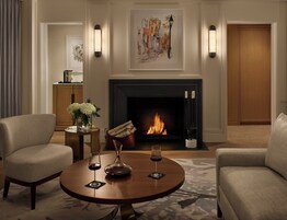A cozy living room with a fireplace, two armchairs, a coffee table with wine glasses, and a painting above the fireplace.