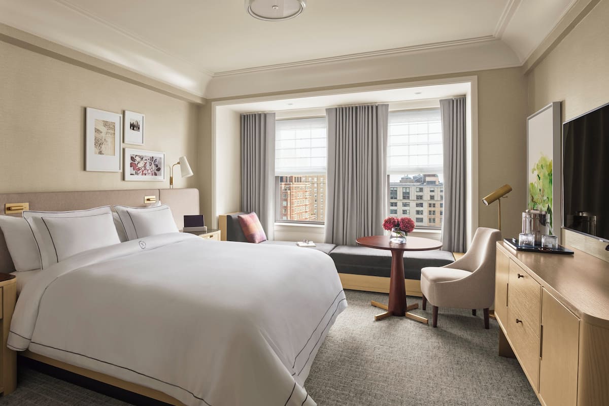 A hotel room with a large bed, a seating area with a view, a small table with a vase, and a TV on the right.