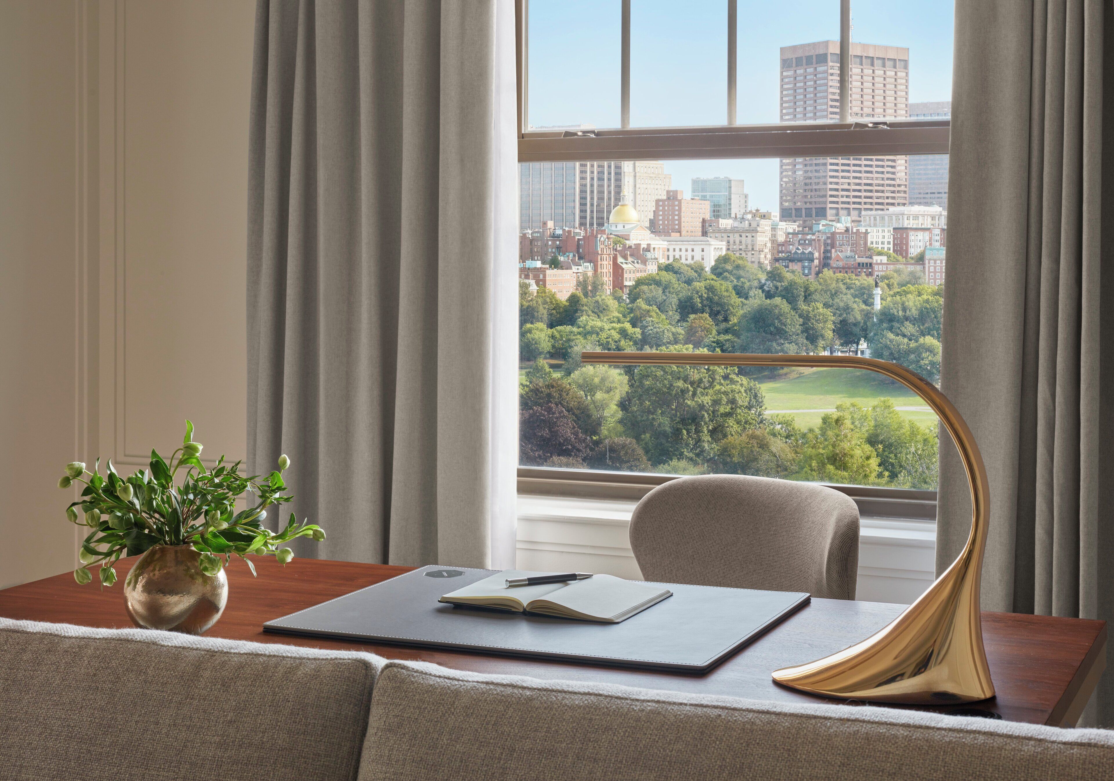 A room with a view of a cityscape through a window, a desk with a laptop, a plant, and a lamp.