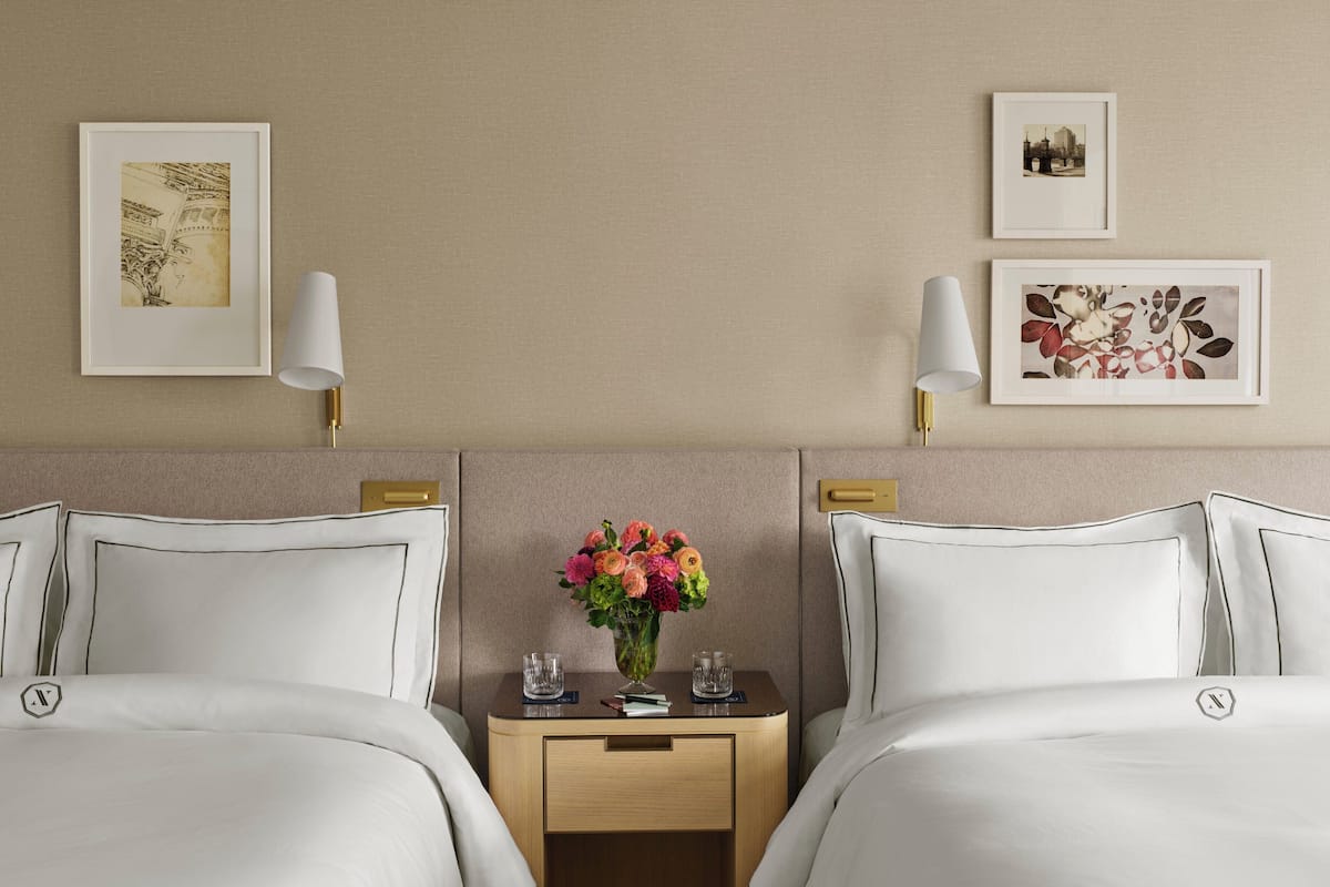 A hotel room with two beds, a nightstand with a vase of flowers, and framed pictures on the wall.