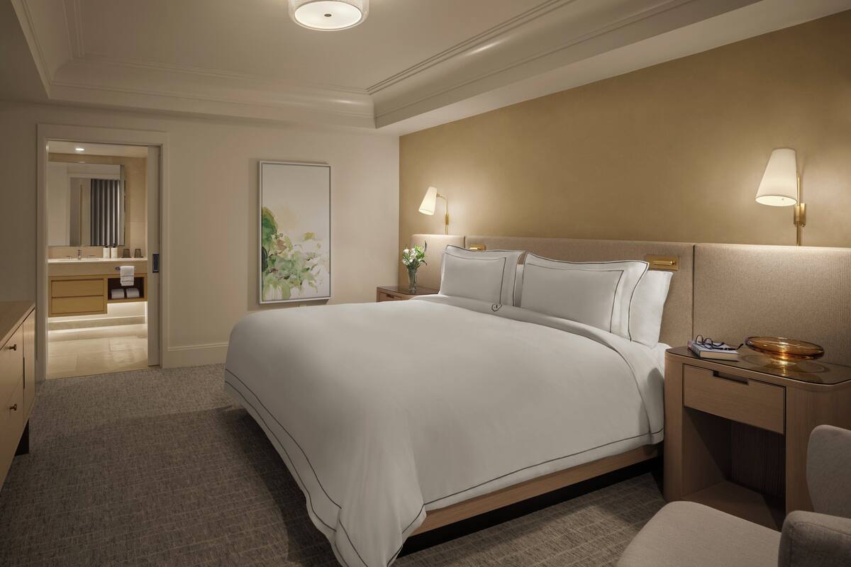 A hotel room with a large bed, bedside tables, a nightstand, a chair, and a bathroom visible through an open door.