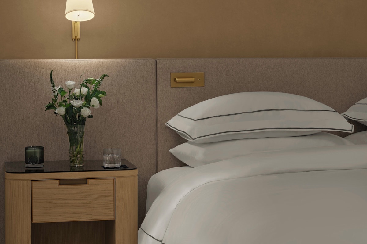 A neatly made bed with white linens, a bedside table with a lamp, a vase with flowers, and a glass of water.