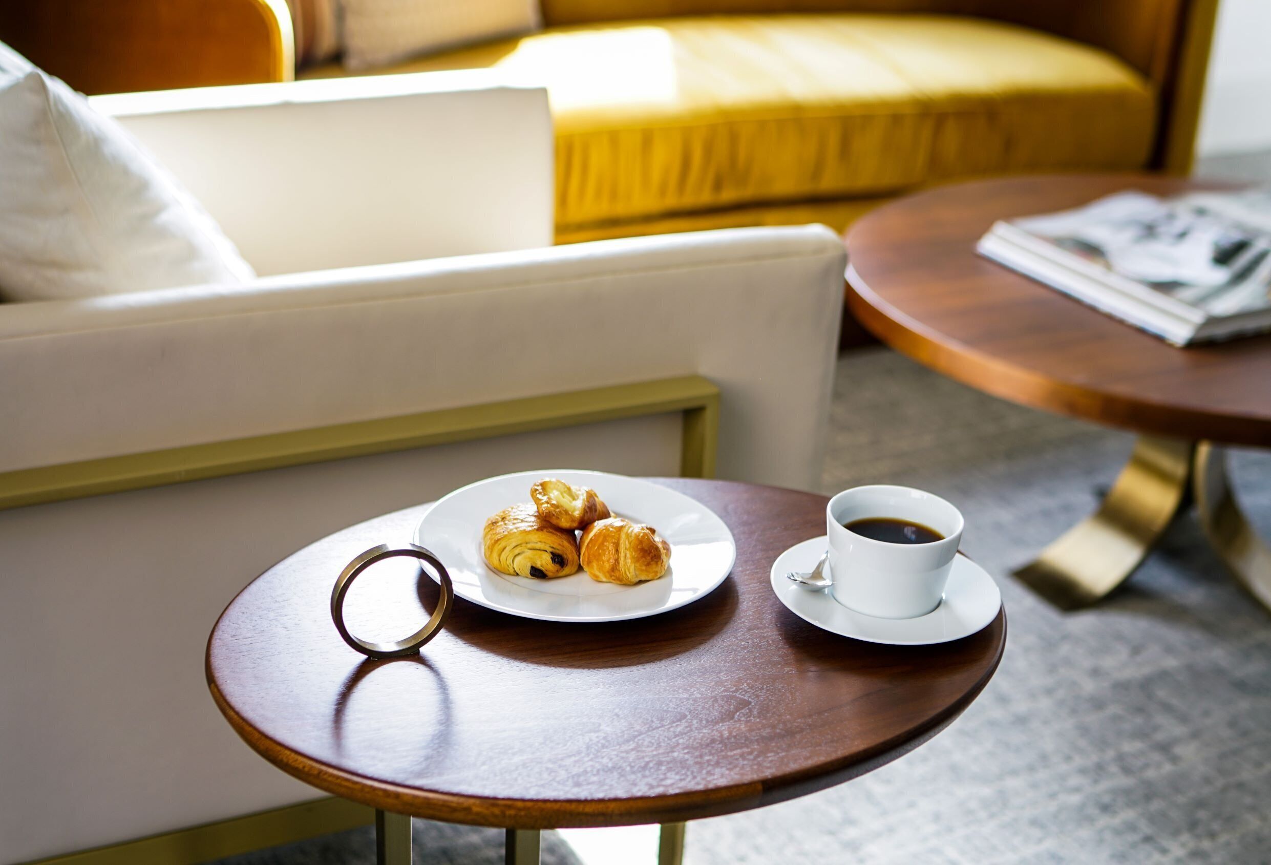 A small round wooden table with a plate of croissants and a cup of coffee, a white sofa, and a yellow armchair in the background.