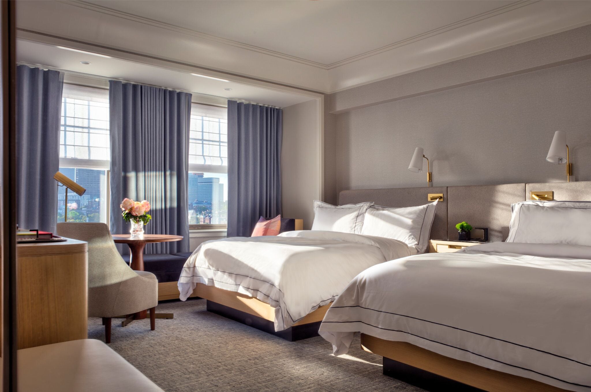 A hotel room with two beds, a desk, a chair, a small table with a vase of flowers, and a view of the city through large windows.