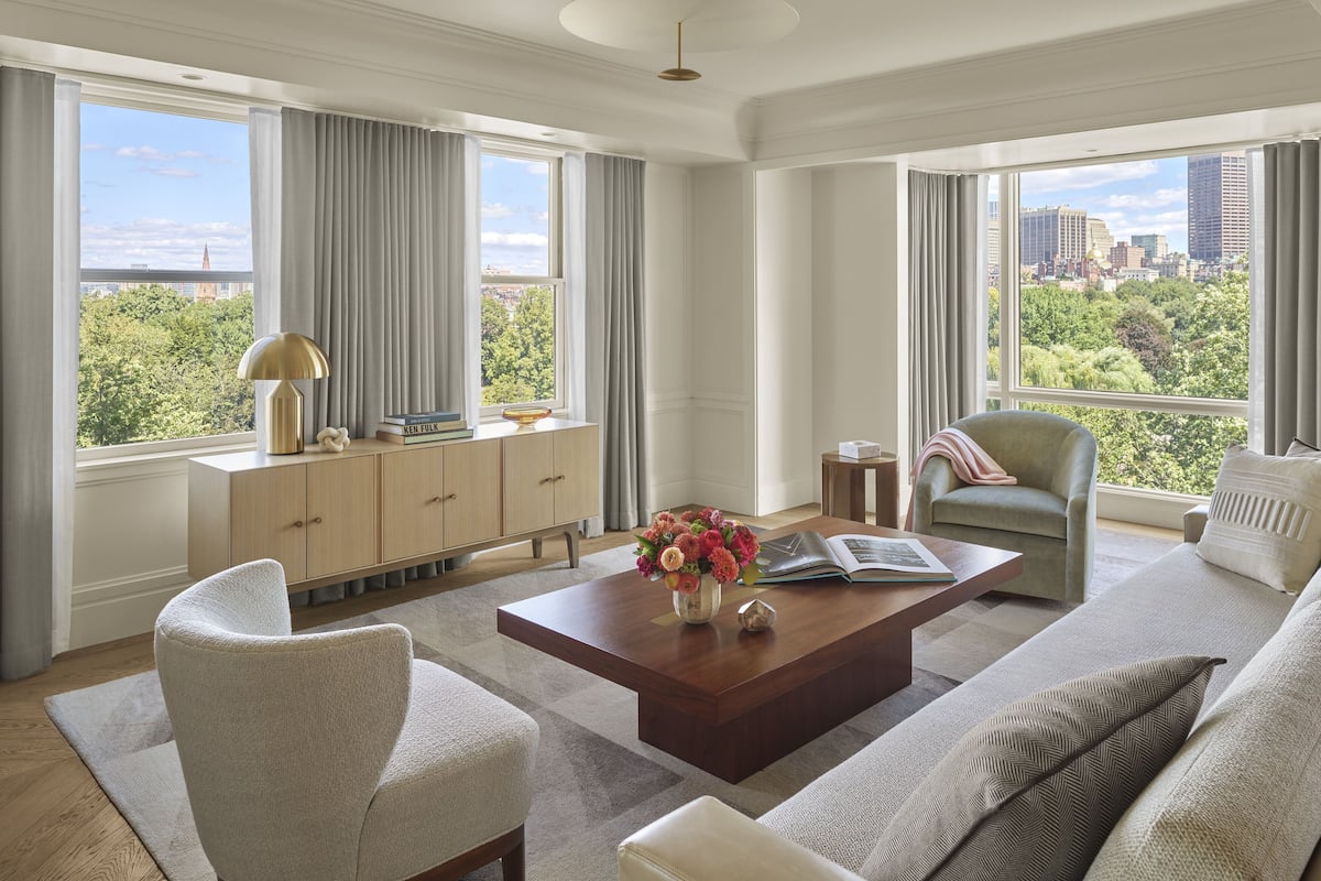 A spacious living room with a large window offering a city view, a wooden coffee table with books and a vase of flowers, and a comfortable seating area with a sofa and armchairs.