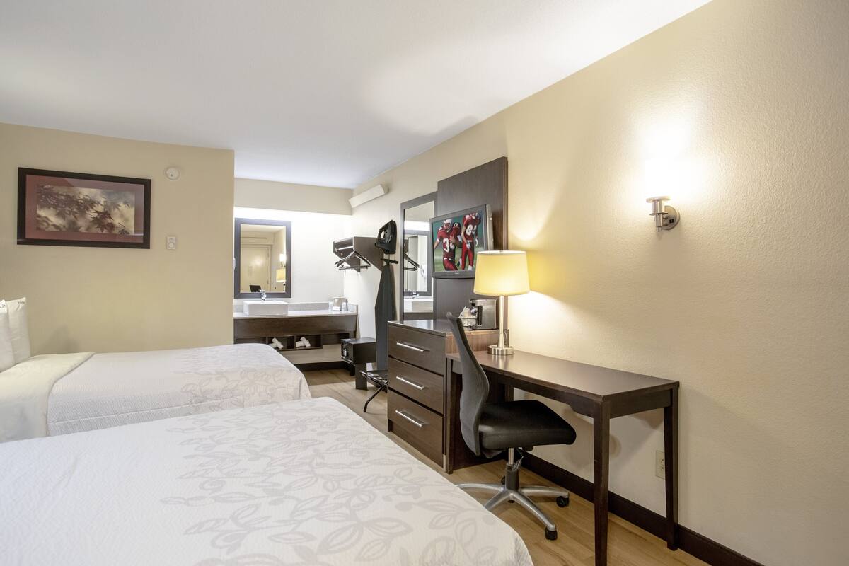 A hotel room with two beds, a desk, a chair, a desk lamp, a mirror, and a wall-mounted light fixture.