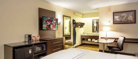 Habitación de hotel con cama, escritorio, silla, televisor, cafetera y espejo.