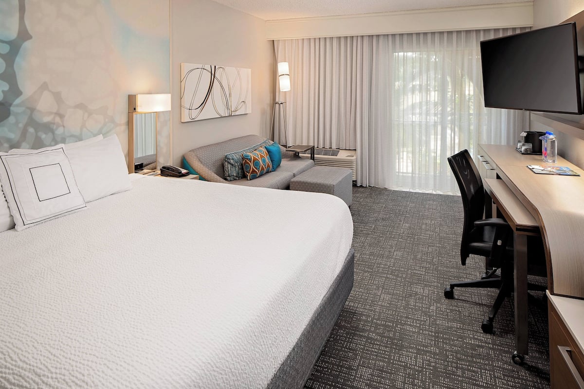 A hotel room with a large bed, a desk with a chair, a flat-screen TV, a lamp, and a window with curtains.