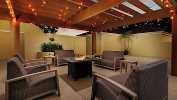 Un patio al aire libre con mobiliario de mimbre, una chimenea central y luces colgantes.