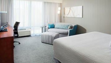 A hotel room with a bed, a desk with a computer, a chair, a lamp, and a large window with curtains.