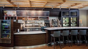 A bar area with a wooden counter, bar stools, a refrigerated display case, and a menu board. - Courtyard by Marriott Fort Lauderdale North/Cypress Creek (Fort Lauderdale)