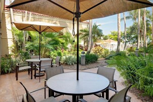A patio area with round tables and chairs, surrounded by lush greenery and palm trees.
