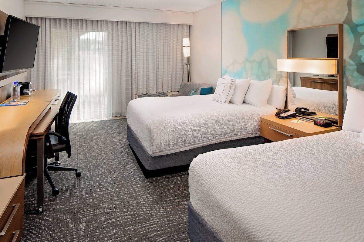 A hotel room with two beds, a desk, a chair, a TV, and a large window with curtains.