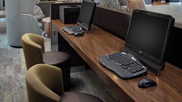Un espace de bureau moderne avec un grand bureau en bois, deux ordinateurs portables, un ordinateur de bureau et une imprimante. On y trouve deux chaises ergonomiques et une petite table ronde avec une lampe.