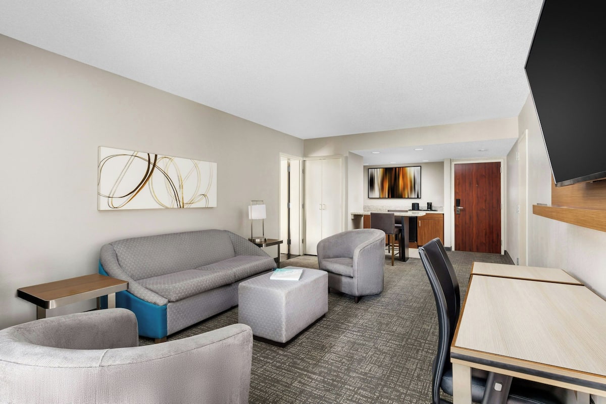 A modern hotel room with a grey sofa, two armchairs, a small table, and a TV.
