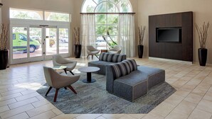 A spacious living area with a large window, a sofa set, a coffee table, and a flat-screen TV mounted on the wall. - Courtyard by Marriott Fort Lauderdale North/Cypress Creek (Fort Lauderdale)