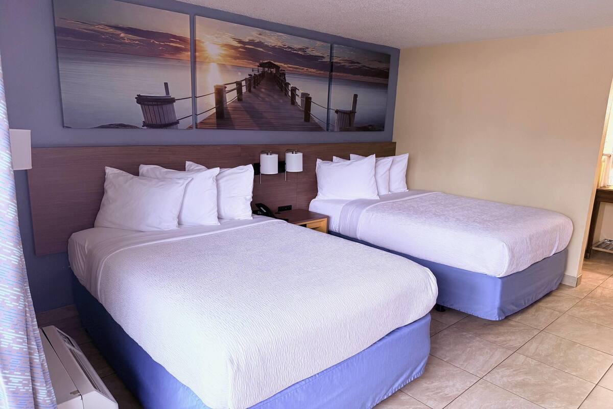 A hotel room with two beds, a nightstand, a wall-mounted lamp, and a sunset painting.