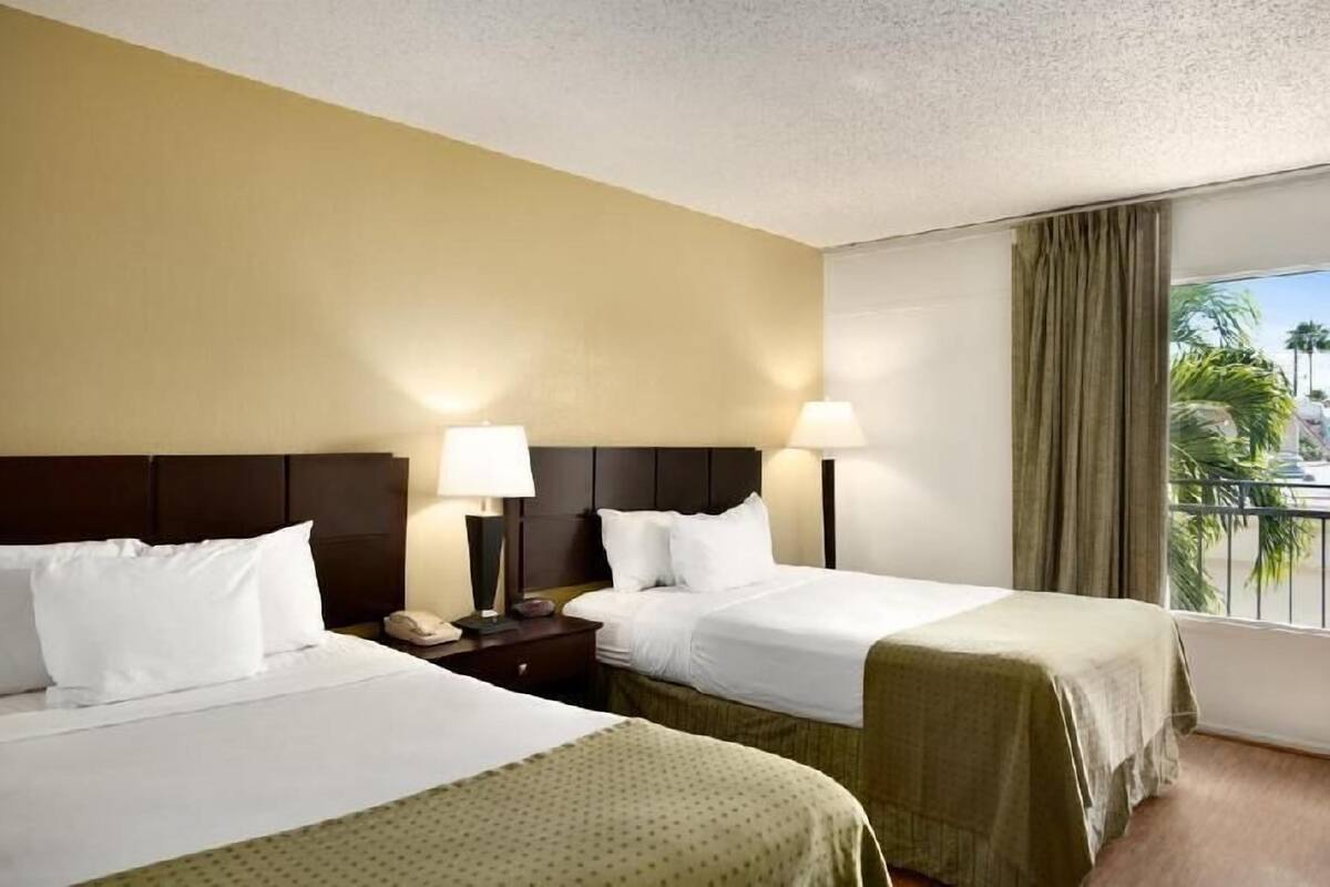 A hotel room with two beds, a nightstand, a lamp, and a view of palm trees.