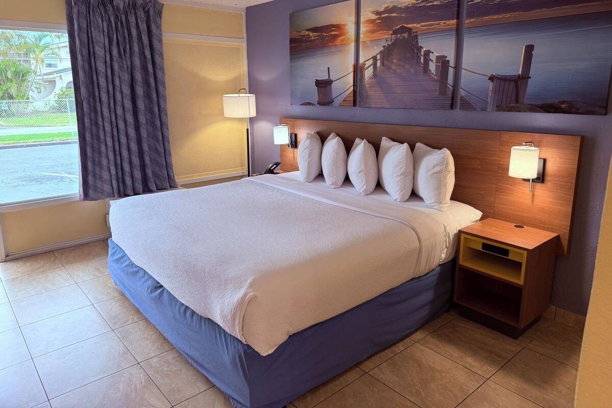 A hotel room with a large bed, a nightstand, a lamp, and a painting of a pier on the wall.