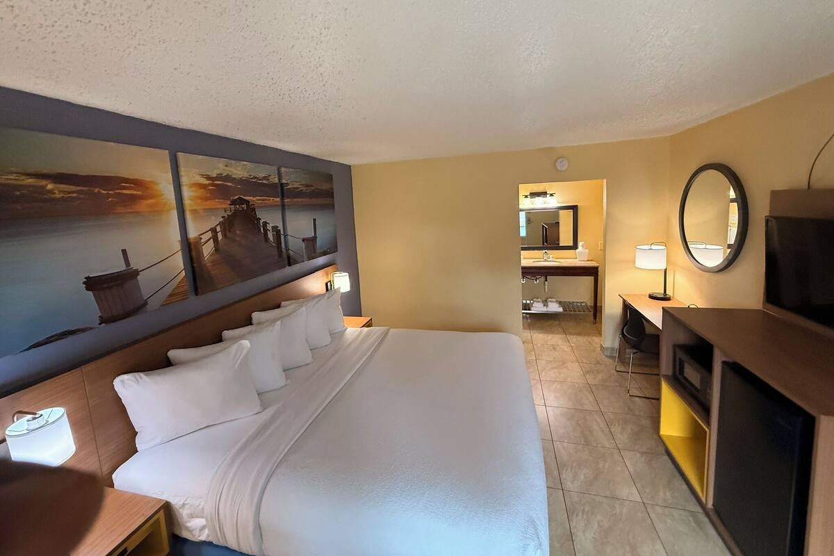 A hotel room with a large bed, a desk with a lamp, a TV, a bathroom with a mirror, and artwork on the wall.