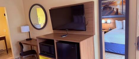 A hotel room with a flat-screen TV, a compact refrigerator, a desk with a chair, a mirror, and a bed with a view of a sunset over a pier.