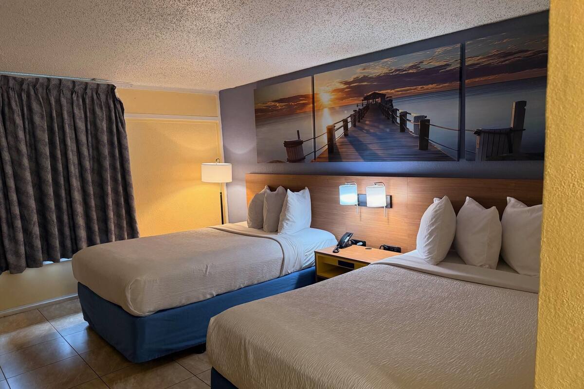 A hotel room with two beds, a nightstand, a lamp, and a sunset painting on the wall.