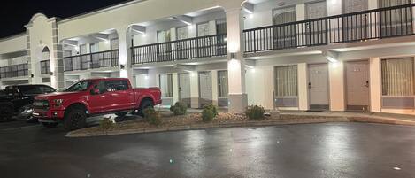 Un hotel de varios pisos con balcones, un estacionamiento y una camioneta roja estacionada al frente.