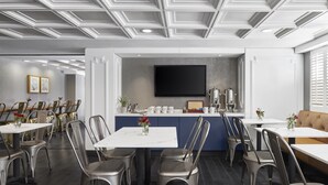 A modern dining area with multiple tables and chairs, a central counter with a flat-screen TV, and a sofa in the background. - Capitol Hill Hotel (Washington)