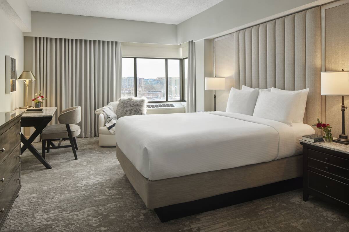 A hotel room with a large bed, a desk, a chair, and a view of the city through the window.
